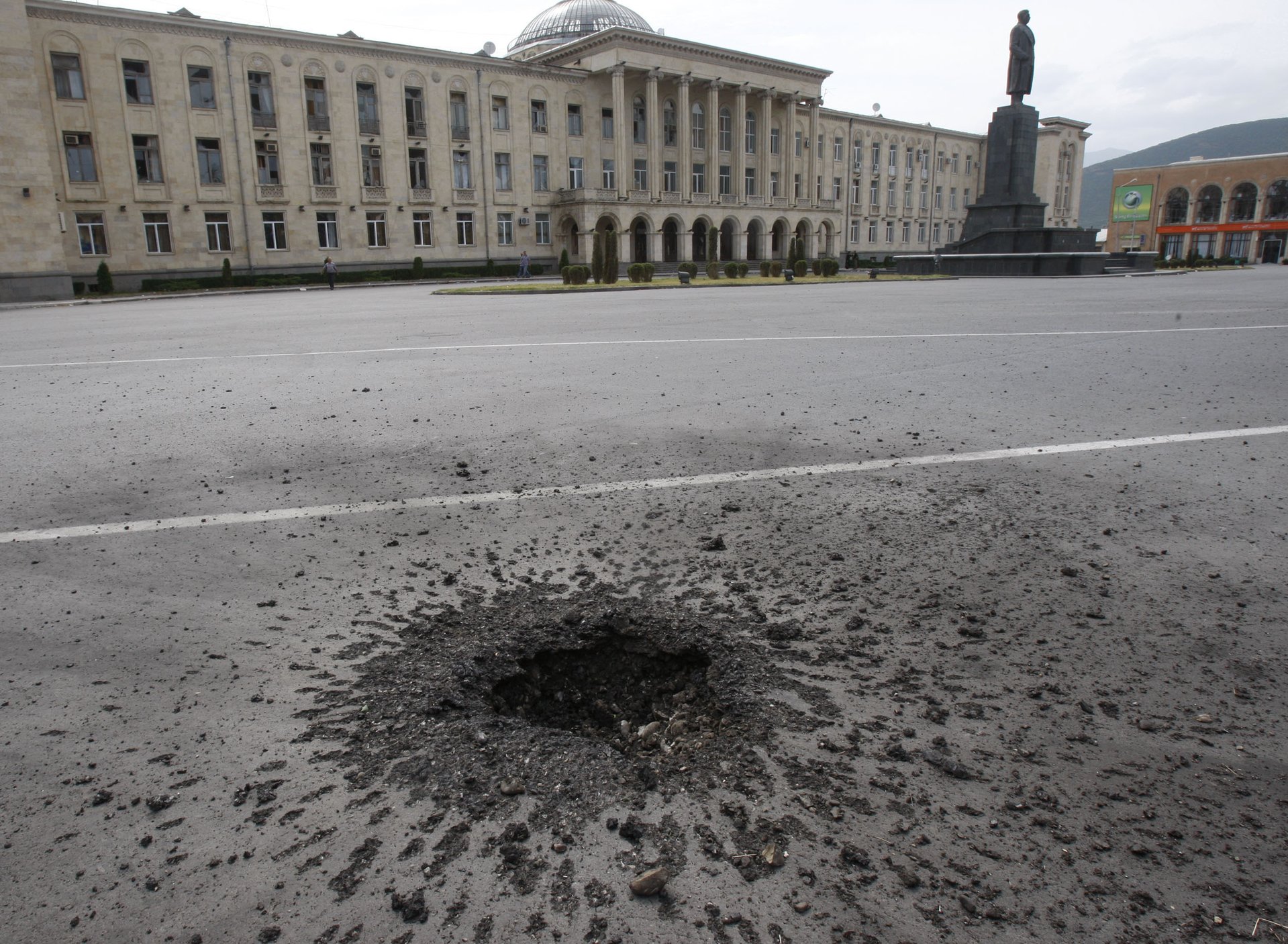 След от взрыва кассетного боеприпаса ракеты «Искандер» на центральной площади Гори с памятником Иосифу Сталину на фоне. Удар унес жизни как минимум шести человек