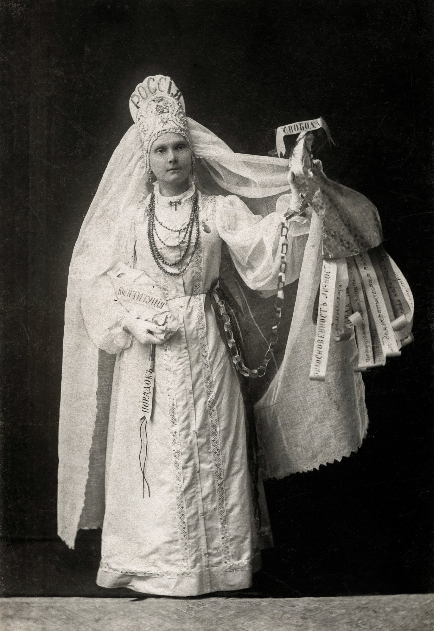 Неизвестная девушка в русском народном костюме. На кокошнике написано «Россия». В правой руке «Россия» держит запеленатого младенца, перевязанного лентой «Конституция». В левой руке рог изобилия с надписью «Свобода», из которого аллегорическим образом высыпаются дары Свободы: неприкосновенность личности, свободомыслие и т. д. Неизвестная мастерская, предположительно 1917 год